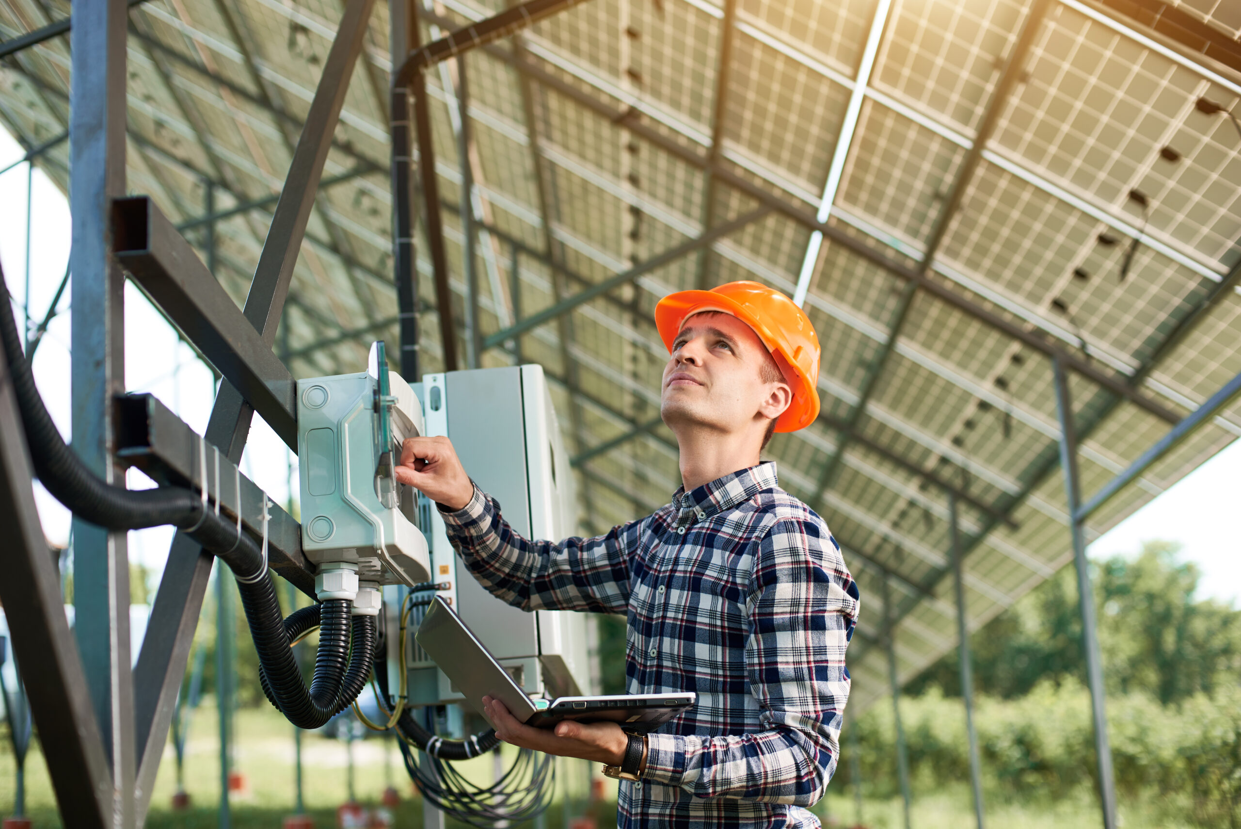 Male engineer in helmet and with laptop pushing button at the electrical equipment on solar power station, looking up. Science technology. Solar panels. Global warming. Green energy.